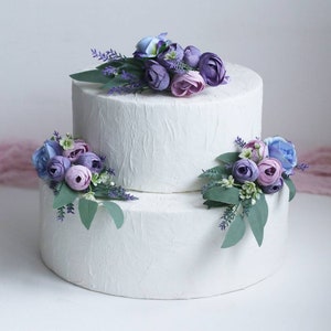 May include: Two-tiered white wedding cake decorated with purple, lavender, and blue artificial flowers.