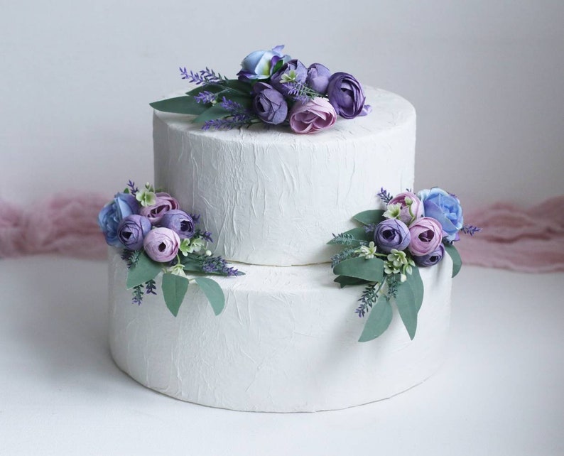 May include: Two-tiered white wedding cake decorated with purple, lavender, and blue artificial flowers.