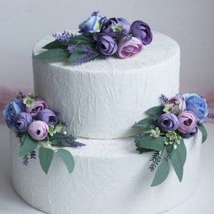 May include: Two-tiered white wedding cake decorated with purple, lavender, and blue flowers. The cake is on a white surface.