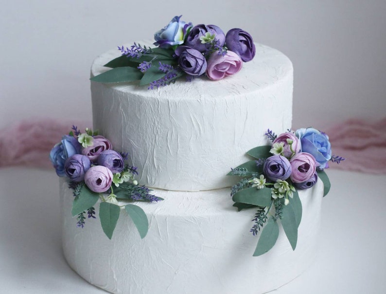 May include: Two-tiered white wedding cake decorated with purple, lavender, and blue flowers. The cake is on a white surface.