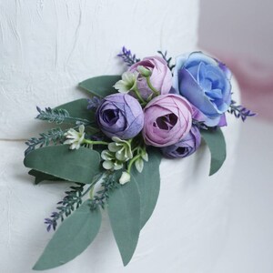 May include: A white wedding cake decorated with a bouquet of purple and blue artificial flowers, including roses and eucalyptus leaves.