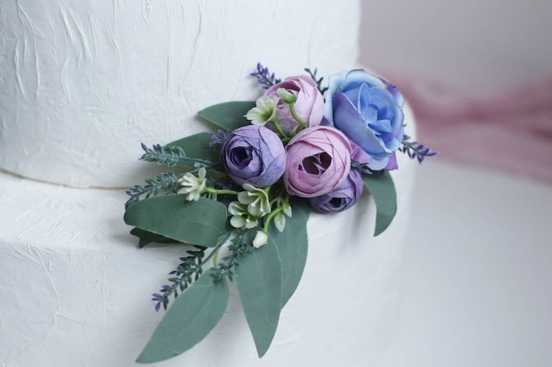 May include: A white wedding cake decorated with a bouquet of purple and blue artificial flowers, including roses and eucalyptus leaves.