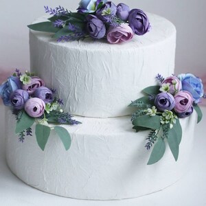 May include: Two-tiered white wedding cake decorated with purple and blue flowers. The cake is frosted with a textured finish.