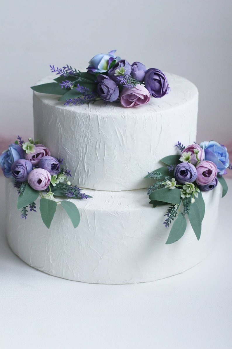 May include: Two-tiered white wedding cake decorated with purple and blue flowers. The cake is frosted with a textured finish.