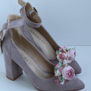 May include: A pair of mauve-colored high-heeled shoes with ankle straps and floral embellishments. The heels are block style, and the shoes have pointed toes. The floral accents are pink and white.