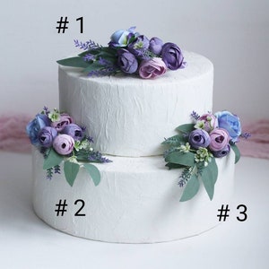 May include: Two-tiered white cake decorated with purple, blue, and lavender artificial flowers. The cake is labelled #1, #2, and #3.