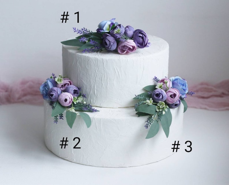 May include: Two-tiered white cake decorated with purple, blue, and lavender artificial flowers. The cake is labelled #1, #2, and #3.