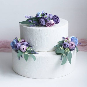 May include: Two-tiered white wedding cake decorated with purple, lavender, and blue flowers.