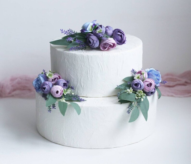 May include: Two-tiered white wedding cake decorated with purple, lavender, and blue flowers.
