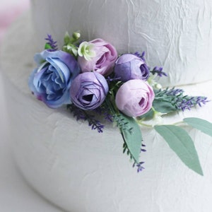 May include: A white wedding cake decorated with a bouquet of purple and blue artificial flowers. The flowers include roses and lavender.