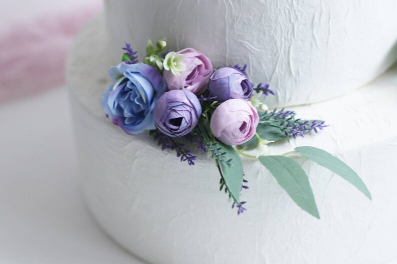 May include: A white wedding cake decorated with a bouquet of purple and blue artificial flowers. The flowers include roses and lavender.