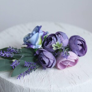 May include: A white cake decorated with a purple and blue flower arrangement. The flowers are made of silk and include lavender, roses, and other blooms.