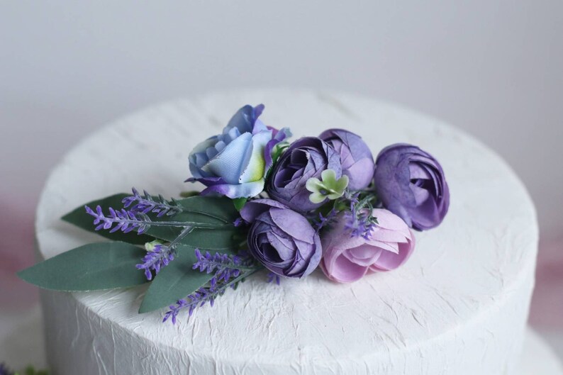 May include: A white cake decorated with a purple and blue flower arrangement. The flowers are made of silk and include lavender, roses, and other blooms.