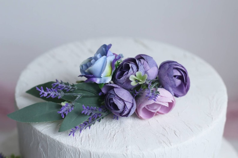 May include: A white cake decorated with a purple and blue flower arrangement. The flowers are made of silk and include lavender, roses, and other blooms.