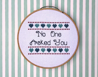 May include: A white embroidery hoop with a cross-stitch design that says "No One Asked You" surrounded by rows of red and blue hearts.