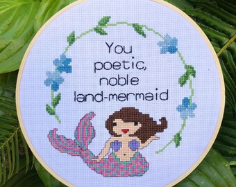May include: A white embroidery hoop with a cross-stitch design of a mermaid with brown hair and a pink tail. The text "You poetic, noble land-mermaid" is stitched in black thread.