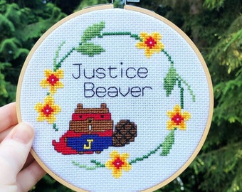 May include: A white embroidery hoop with a cross-stitch design of a brown beaver wearing a red and blue superhero costume with a yellow 'J' on the chest. The beaver is surrounded by yellow flowers and green leaves. The text 'Justice Beaver' is stitched in black thread above the beaver.