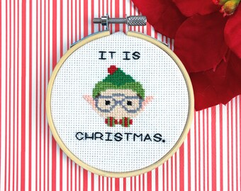 May include: A white embroidery hoop with a cross-stitch design of an elf wearing glasses and a green hat. The text "IT IS CHRISTMAS." is stitched below the elf.