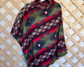May include: A patterned poncho in shades of olive green, red, and gray, with a geometric design. The poncho features a cowl neck and decorative silver buttons on the shoulder. The garment is hanging on a black hanger.