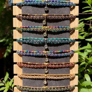 May include: A selection of colourful macrame bracelets on display. The bracelets, in shades of blue, brown, green, and gold, are adorned with beads and charms. They are presented on a wooden stand with a dark grey backing, showcasing the intricate knotting of each piece.