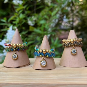 Pode incluir: Três anéis de macramé feitos à mão em verde, azul e dourado, cada um adornado com contas douradas e uma gema transparente pendurada. Os anéis são exibidos em pequenos cones marrons, em um fundo verde e branco desfocado.