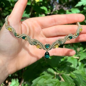 Op de afbeelding: Een handgemaakte macramé ketting met groene en gele kristallen kralen en een druppelvormige hanger. De ketting is gemaakt met een beige koord en goudkleurige accenten. Het ontwerp wordt in een hand gehouden tegen een achtergrond van groen gebladerte.
