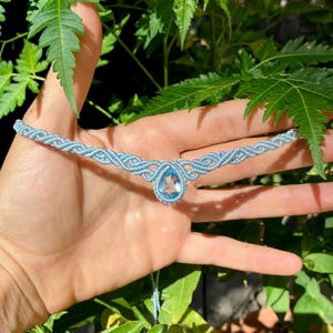 May include: A light blue macrame choker necklace featuring a central teardrop-shaped faceted crystal pendant. The band is crafted from intricate knotted cord, creating a repeating geometric pattern that extends outward from the crystal center.