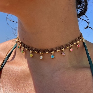 May include: A brown braided choker necklace with dangling colorful gemstones. The necklace features a variety of colors, including green, yellow, blue, pink, and clear.