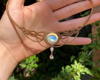 Puede incluir: Collar de macramé hecho a mano con un colgante central de piedra lunar y un pequeño cristal colgante. El collar está hecho con cordón de color beige y detalles de cadena dorada. El diseño incluye intrincados nudos y un estilo bohemio delicado.