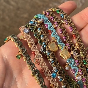 May include: A collection of handmade macrame bracelets in various colors, adorned with beads and charms. The bracelets feature gold, silver, and gemstone accents, including stars, shells, and crystals. The bracelets are displayed on a hand.