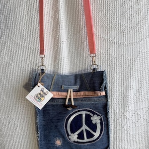 May include: A denim crossbody bag with a pink strap and a peace sign applique. The bag features a white peace sign and flower design on a dark blue denim background. The bag has a drawstring closure and a tag that reads "Peace Sign Girls".