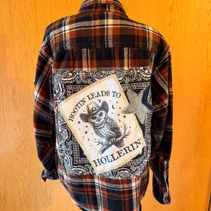May include: A long-sleeve button-down flannel shirt in a brown, black, and white plaid pattern. The back features a large white fabric patch with a black graphic of an owl wearing a cowboy hat, the text HOOTIN LEADS TO HOLLERIN, and a denim star.