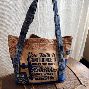 May include: A denim tote bag with a floral patterned top and a tan front panel. The front panel features a cross graphic and the text "Now Faith Is Confidence In What We Hope For And Assurance About What We Do Not See Hebrews 11:1".