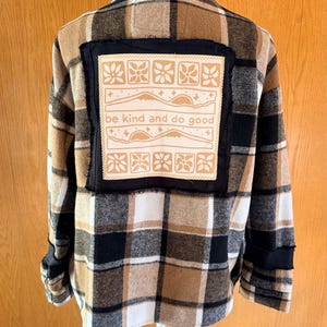 May include: A long-sleeve button-up shirt featuring a brown, black, and white plaid pattern. A large square patch is sewn onto the back, displaying a tan graphic with floral motifs, mountain silhouettes, and the text be kind and do good.