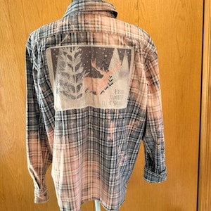 May include: A long-sleeve button-down flannel shirt featuring a bleached plaid pattern in shades of grey, black, and faded peach. The back displays a rectangular graphic of a deer silhouette among pine trees with the text Hello Winter Let It Snow.