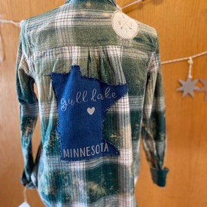 May include: A green and white plaid flannel shirt with a blue denim patch on the back. The patch is shaped like the state of Minnesota and has the text "Gull Lake" and a heart. The shirt also has a small compass patch on the right shoulder.