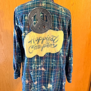 May include: A long-sleeve button-down shirt in a blue, green, and red plaid pattern with a bleached, distressed effect. The back features a dark gray circular peace sign patch and a yellow fabric patch with the text Happiest Camper.