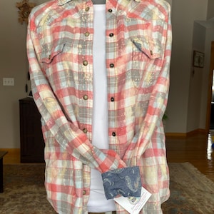May include: A red, white, and gray plaid flannel shirt with a faded, bleached look. The shirt has a button-down front and long sleeves. The cuffs are turned up to reveal a blue paisley pattern.