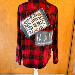 May include: A red and black buffalo plaid long-sleeve shirt featuring a bleached effect. The back displays two rectangular fabric patches with frayed edges. The top patch reads MOST LIKELY TO DECK THE HALLS with colorful Christmas lights.