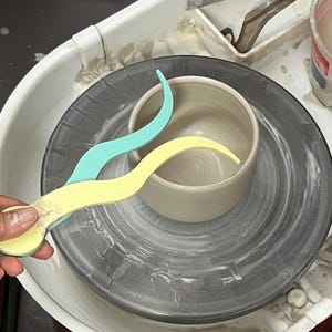 Puede incluir: Un torno de alfarero con un cuenco de cerámica gris y dos herramientas curvas para esculpir arcilla. Una herramienta es de color amarillo claro y la otra azul claro. El cuenco se encuentra sobre una cabeza de rueda gris oscuro.