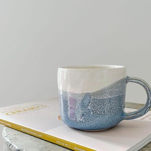 Può includere: Una tazza in ceramica con la parte superiore bianca e la parte inferiore blu strutturata. La tazza ha un manico curvo e poggia su una rivista con la scritta "AUSTRALIAN CERAMICS". Il design della tazza ricorda un acquerello.