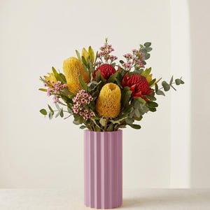 Op de afbeelding: Een levendig bloemstuk in een lavendelkleurige vaas. Het boeket bevat gele, rode en roze bloemen met groen blad. De vaas heeft een geribbeld ontwerp en is ongeveer 25 cm hoog.