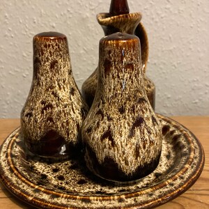 Könnte beinhalten: Ein Keramik-Salz- und Pfefferstreuer-Set mit einer passenden Öl- oder Essigkaraffe. Das Set hat eine braune und cremefarbene, gesprenkelte Glasur. Die Salz- und Pfefferstreuer sind birnenförmig.
