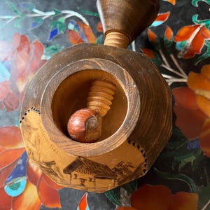 May include: A wooden nutcracker with a screw mechanism, holding a hazelnut. The nutcracker is made of dark wood and has a decorative carved design. The background features a colorful floral pattern.