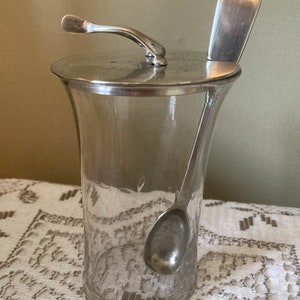 Puede incluir: Un tarro de cristal transparente con tapa y asa plateadas, y una cuchara a juego. El tarro tiene forma acampanada y está sobre un mantel de encaje blanco. La cuchara descansa dentro del tarro y sobre la tapa.