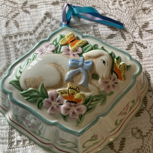 May include: A white ceramic mould shaped like a heart with a raised design of a white bunny with blue ribbon, surrounded by pink and yellow flowers and butterflies. The mould has a blue and green border and a white ribbon attached.