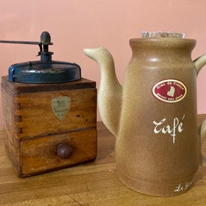 May include: A vintage wooden coffee grinder with a blue metal top and a brown ceramic coffee pot with the text "Cafe" and "Gras de France" on the side.