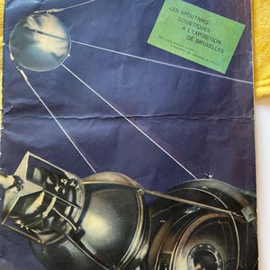 May include: A vintage magazine cover featuring a black and white illustration of a satellite in orbit. The magazine is titled "L'UNION SOVIÉTIQUE" and the text "LES SPOUTNIKS SOVIÉTIQUES A L'EXPOSITION DE BRUXELLES" is visible in a green box.