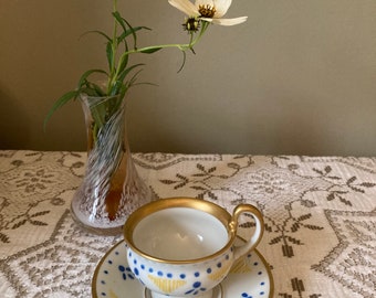 Może przedstawiać: Biały porcelanowy kubek i spodek z pozłacanym rantem i niebiesko-żółtym wzorem kwiatowym. Kubek i spodek znajdują się na białym obrusie z koronki.