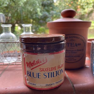 Op de afbeelding: Een vintage pot Webster's Saxelbye Farm Blue Stilton kaas. De pot heeft een bruin deksel en een etiket met rode en blauwe tekst. Op de achtergrond staat een grotere terracotta container.